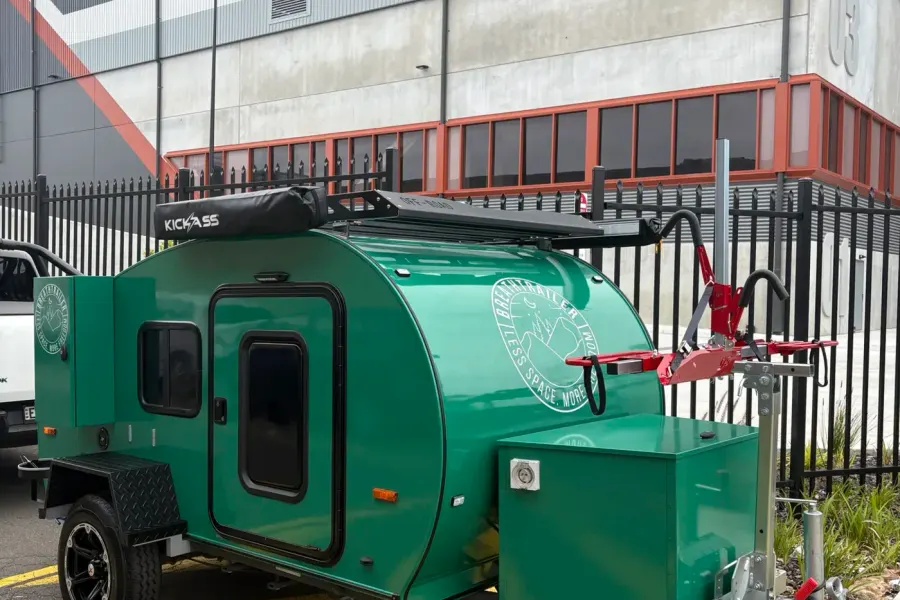 Green compact Breath teardrop with roof rack, drawbar bike rack and front toolbox beside a fence