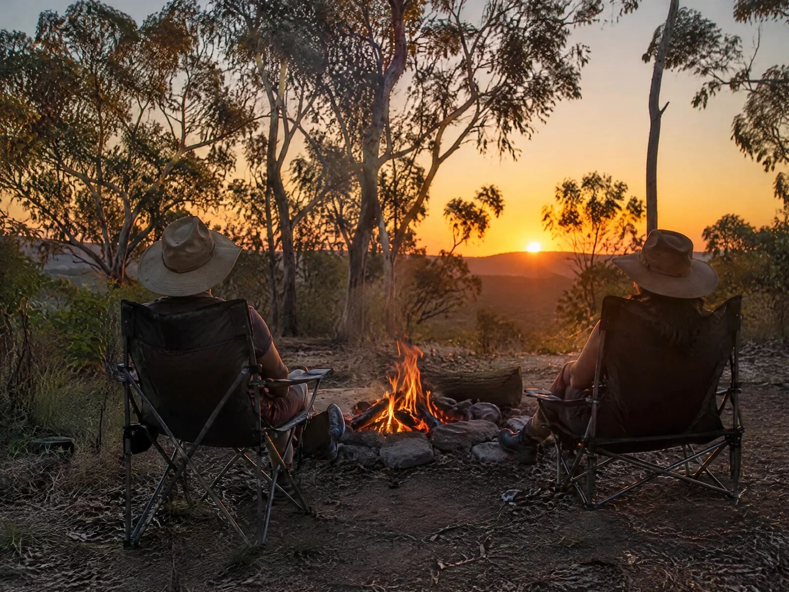 Two people sitting beside a campfire at a bush campsite at sunset with eucalyptus trees behind them