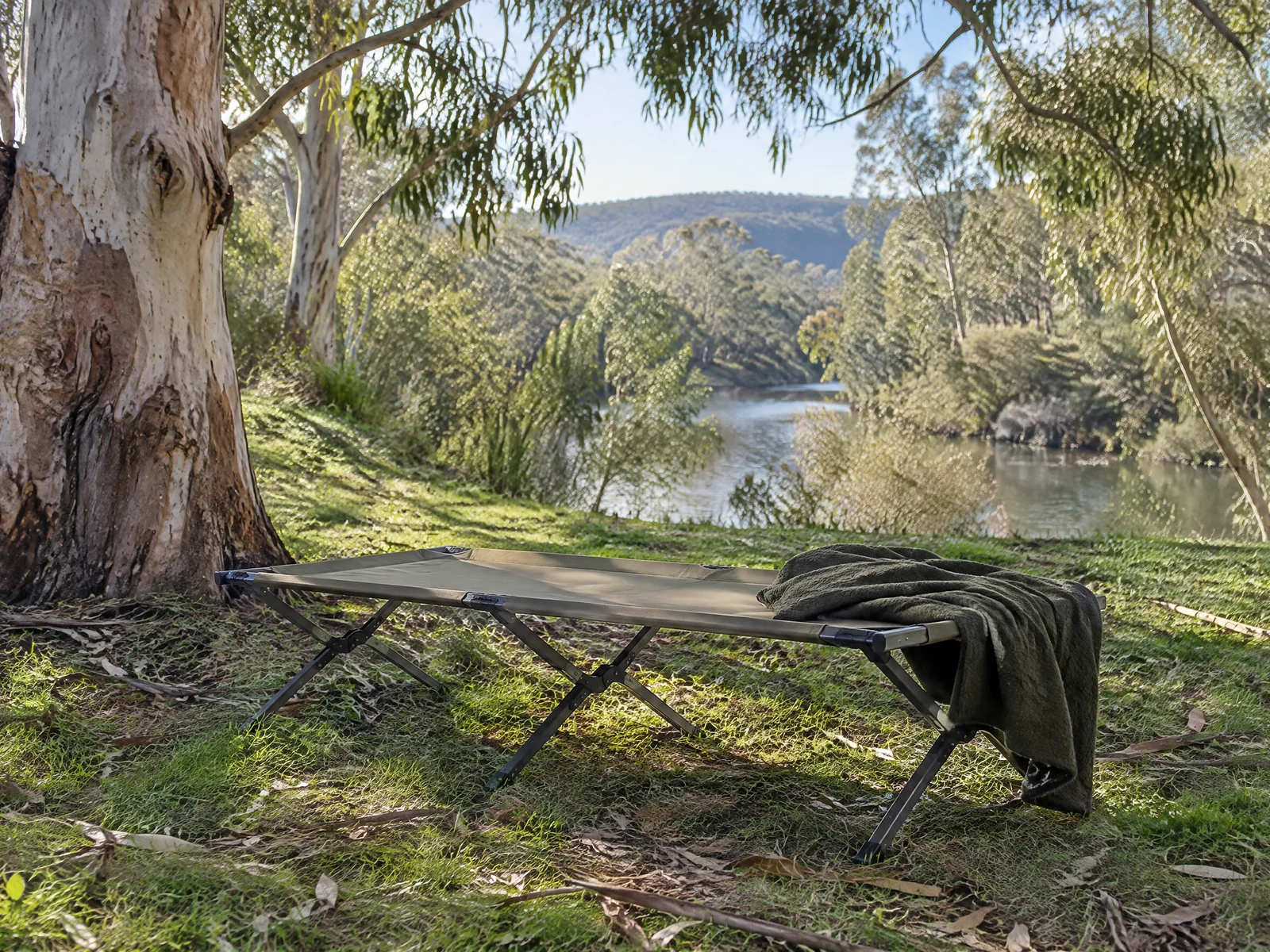A portable camping cot set up in dappled shade beneath eucalyptus trees at a bush campsite