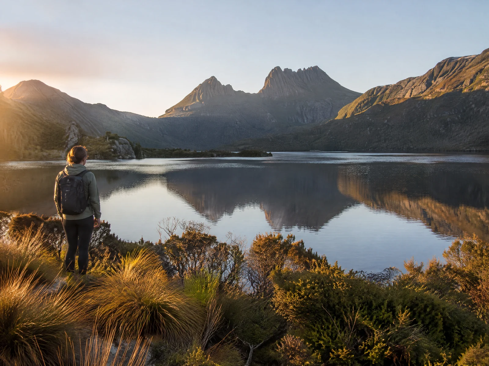 Hiker at Dove Lake with Cradle Mountain's dolerite peak reflected in the still water at golden hour