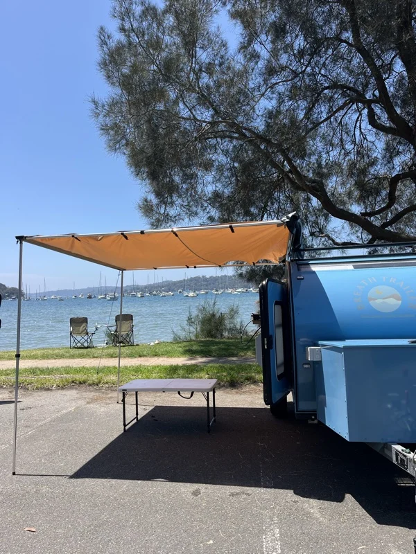 Blue compact Breath teardrop with orange awning, table and chairs overlooking a harbour with yachts