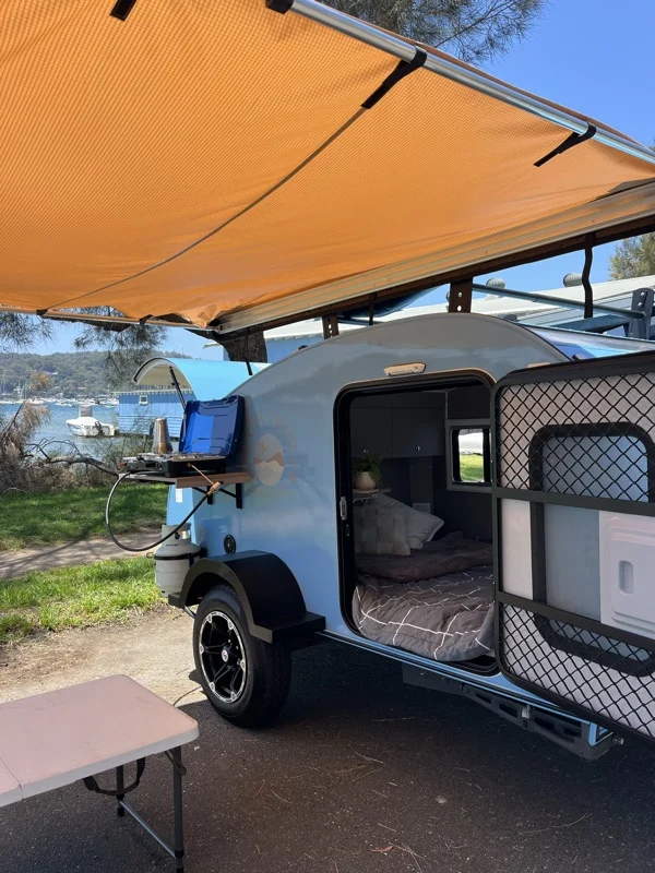 Compact Breath teardrop under an awning by the water, bed made up and stove on the side shelf