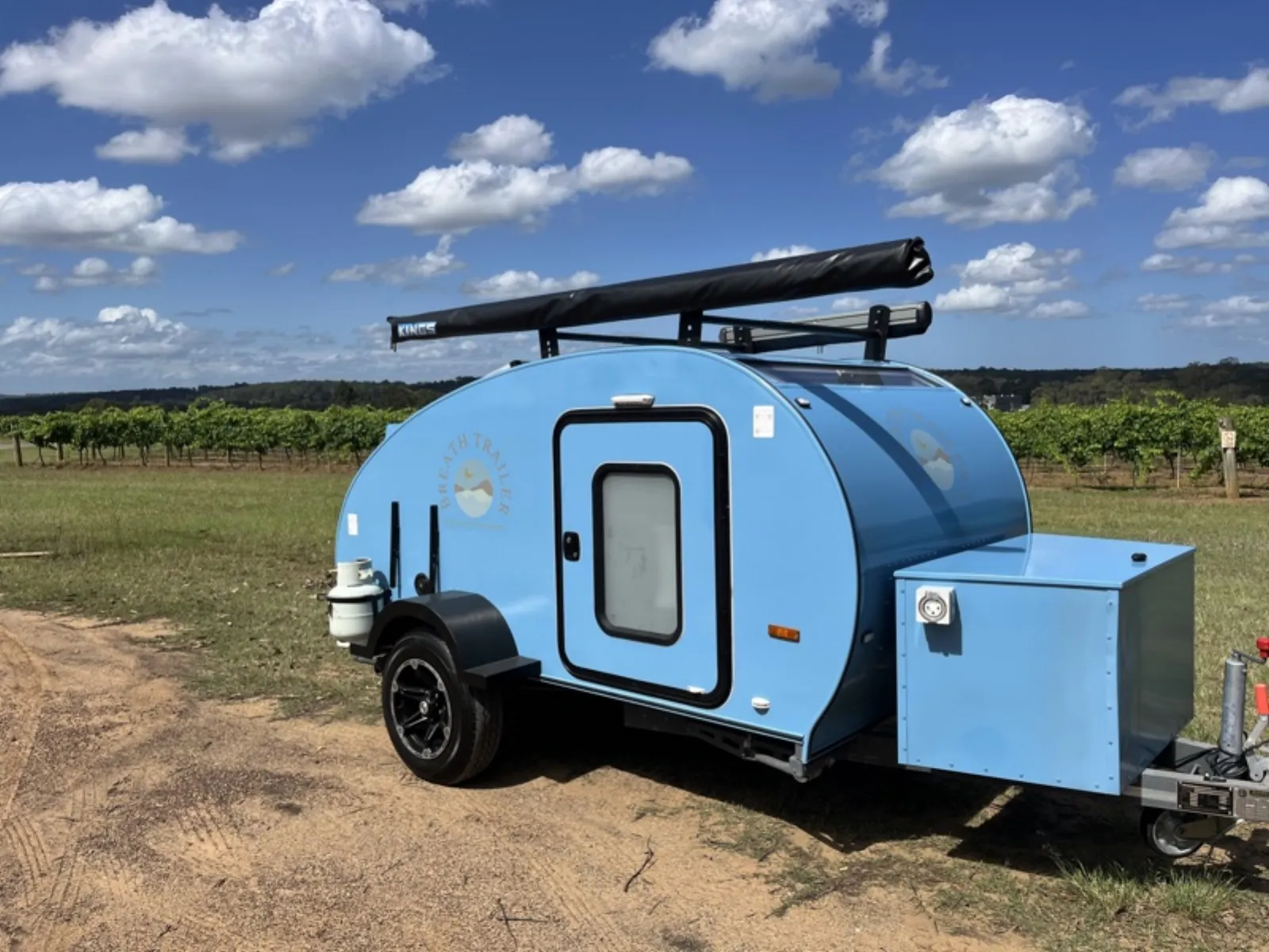 Sky blue compact Breath teardrop with a rolled awning on its roof rack parked beside rows of vineyard vines