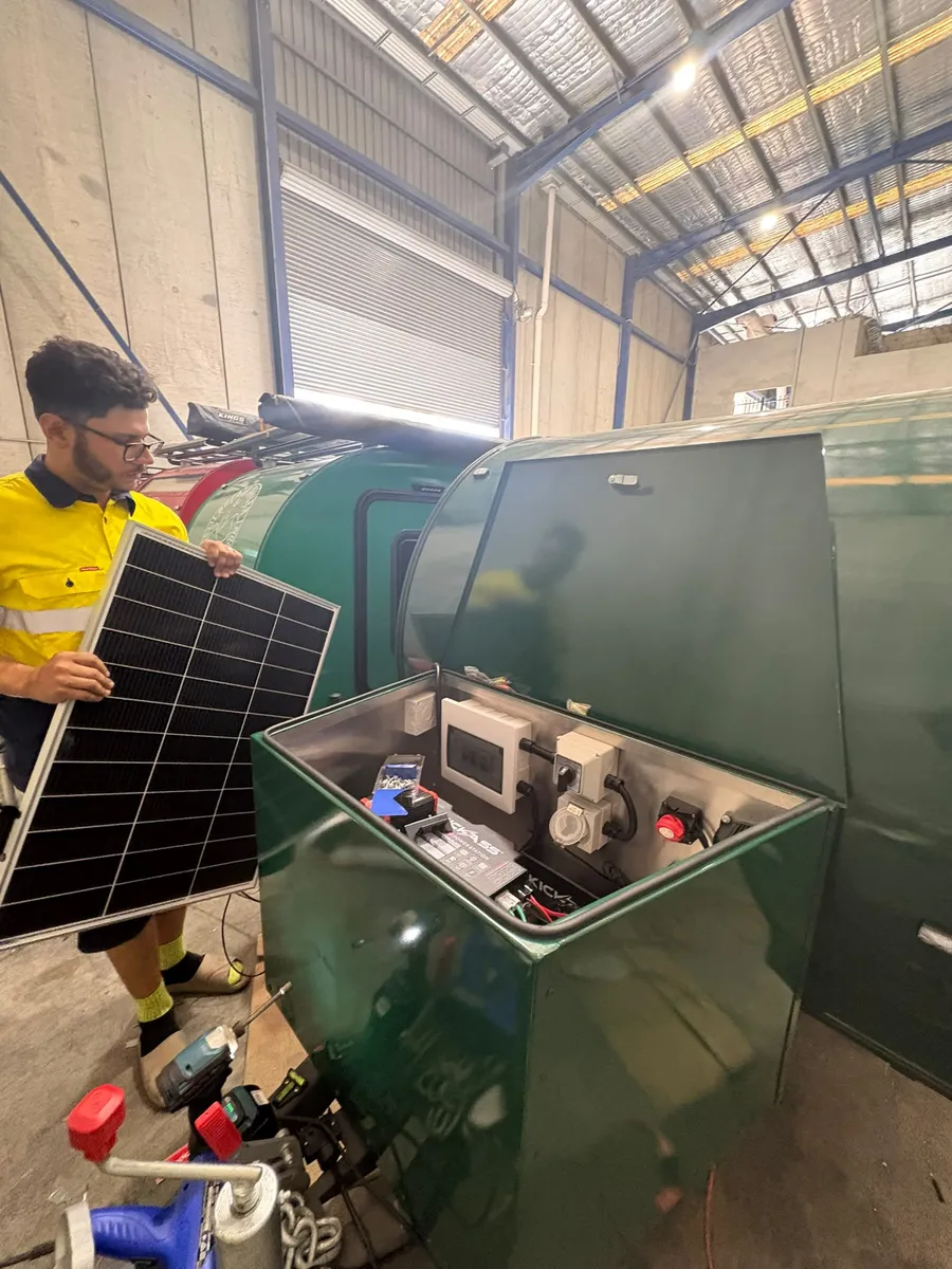 Breath Trailer technician holding a solar panel beside the open battery and electrical box of a green Breath camper in the Sydney workshop